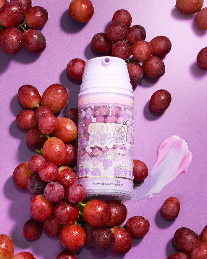 Fialová láhev P. Louise Here I Glow Again Grape Recovery Mask obklopená hrozny na fialovém pozadí