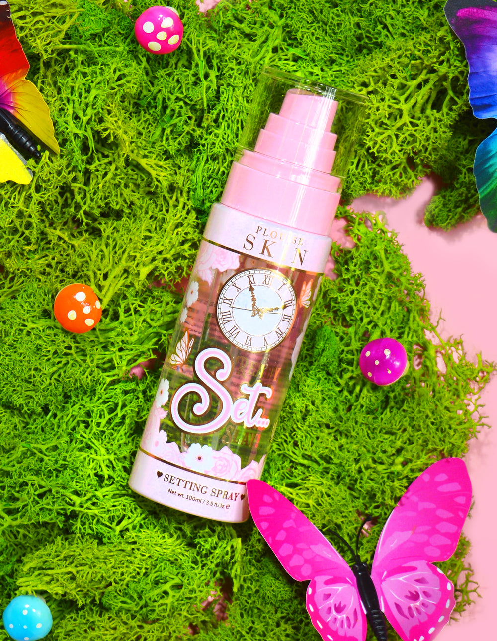 Láhev P. Louise Ready Set Slay Long Lasting Setting Spray obklopená dekorativními prvky včetně barevného umělého mechu, motýlů a hub.