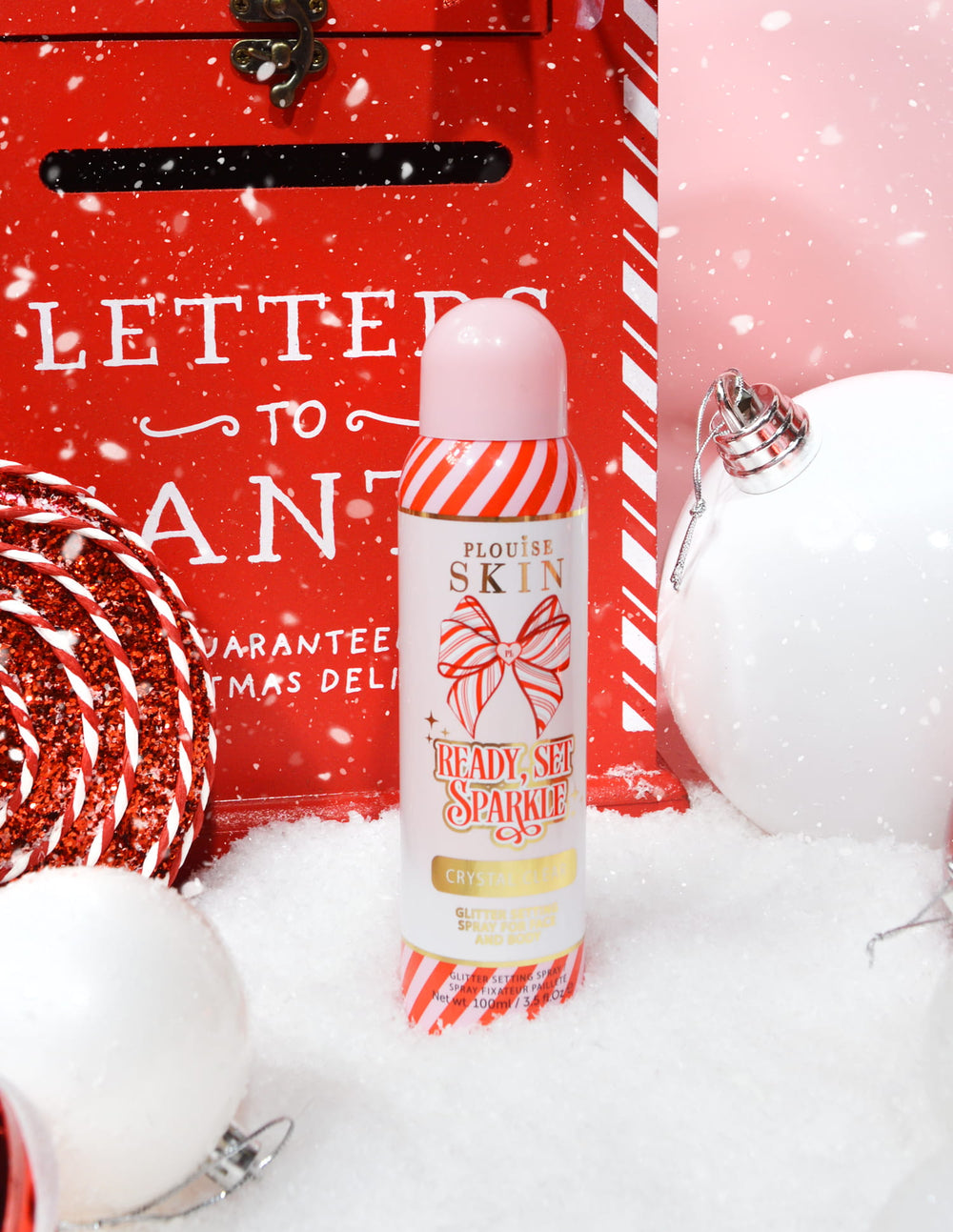 Láhev P. Louise Ready Set Sparkle Christmas Glitter Setting Spray obklopená svátečními dekoracemi, jako je sníh, ozdoby a vánoční poštovní schránka