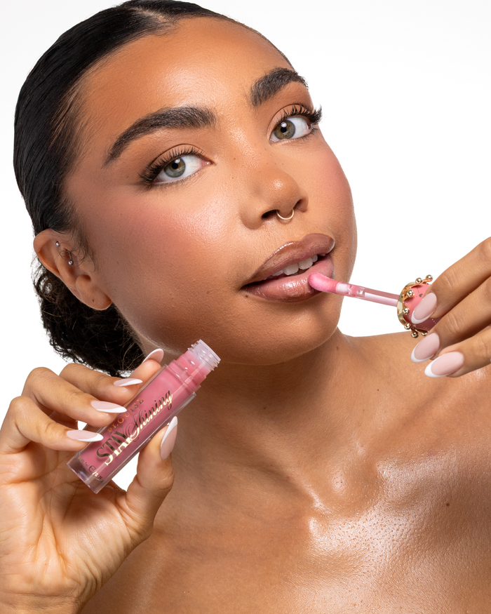 Detail ženy nanášející růžový tekutý produkt na rty P.Louise Stay Shining Lip Oil pomocí aplikátoru s laní nohou, zatímco drží tubu v druhé ruce, na čistém bílém pozadí.