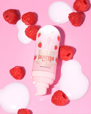 Pink P.Louise Bright Direction Raspberry Brightening Serum skincare product with raspberry design surrounded by raspberries on a pink background