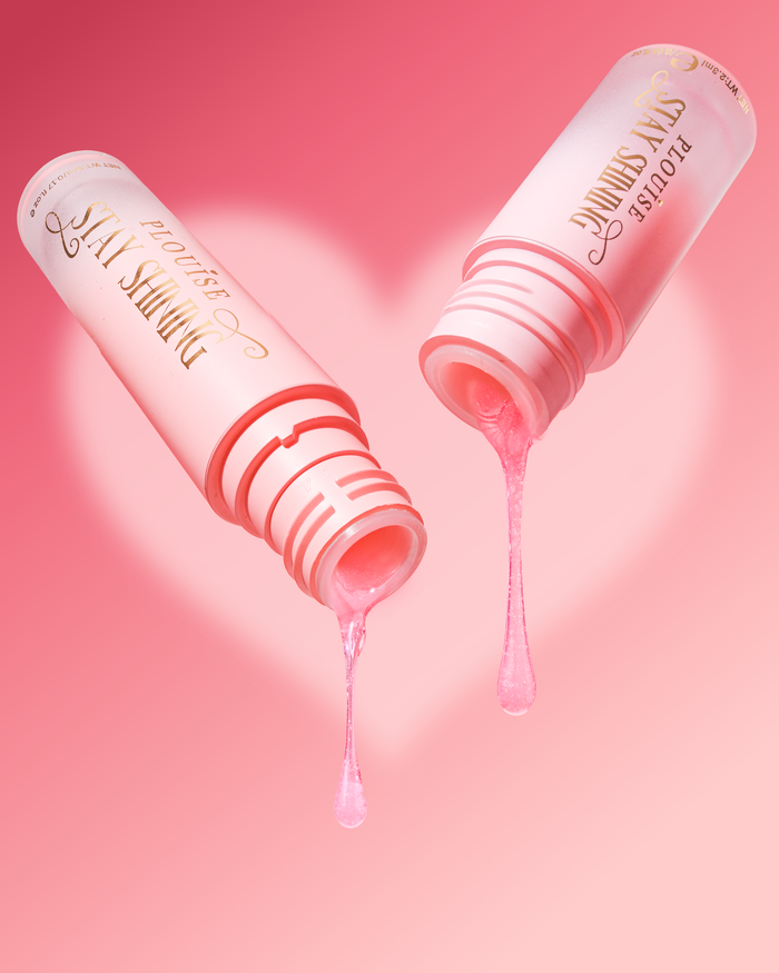 Two pink cosmetic tubes labeled 'STAY SHINING' shown on a pink background with a heart shape behind them. Pink gel is dripping from both open tubes, with one drop falling below and another above.