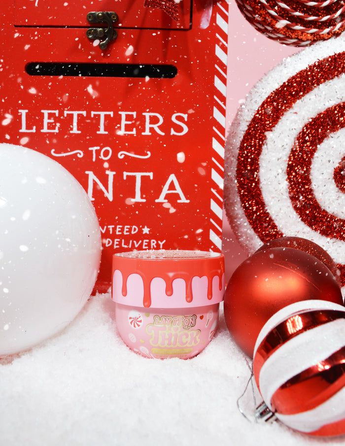 Sklenice P.Louise Lay It On Thick Body Butter ve variantě Frosted Peppermint, vystavená s vánočními ozdobami, včetně vánočních světel a dárků.