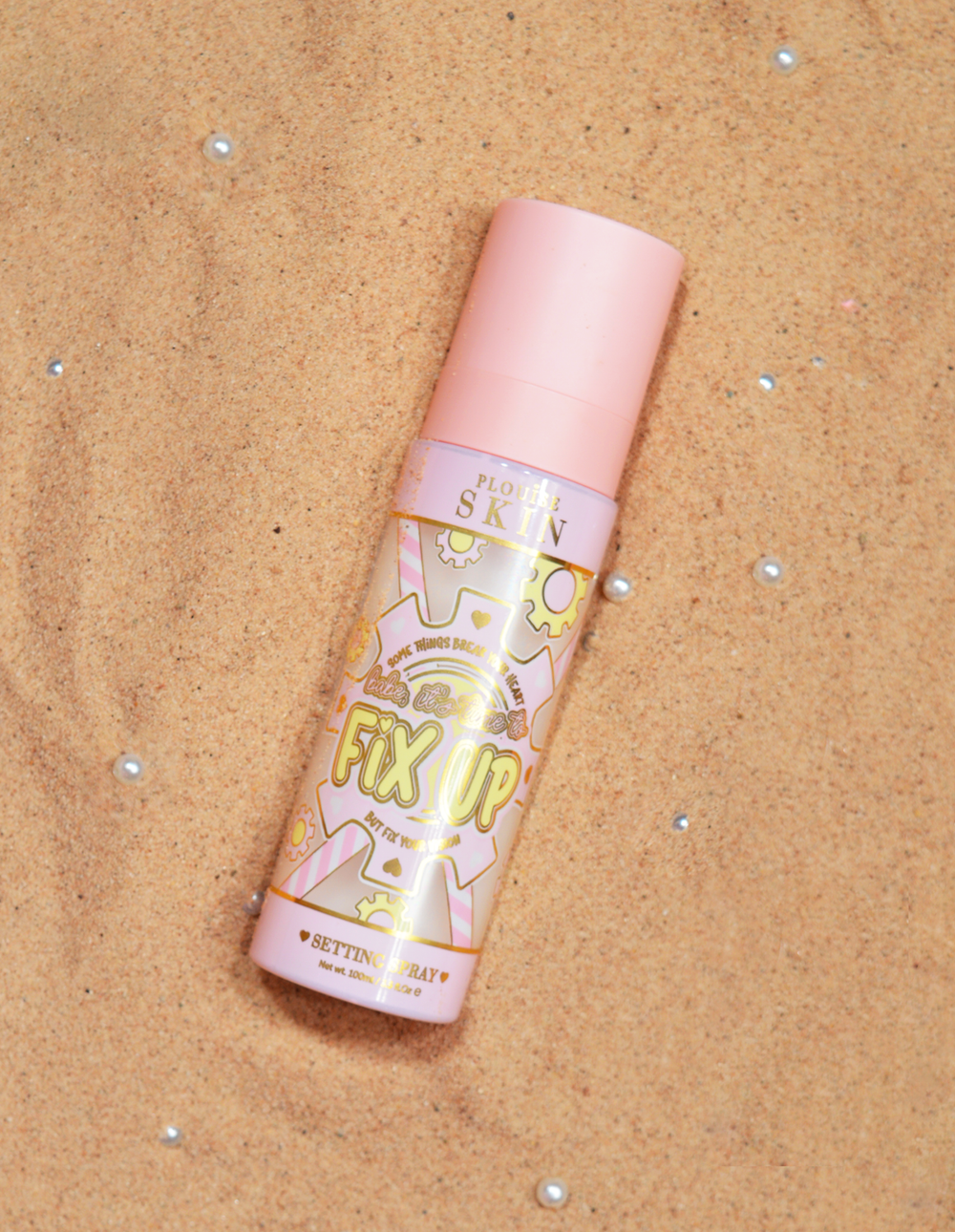 Lahvička P.Louise Fix Up Mattifying Setting Spray s růžovým uzávěrem a ozdobnými vzory na etiketě, vystavená na pískovém povrchu.