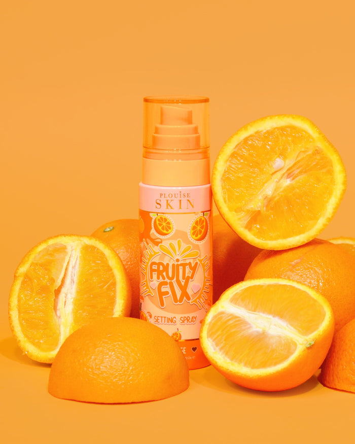 Oranžová lahvička P. Louise Fruity Fix Vitamin C Fixační sprej s oranžovým pozadím obklopená čerstvými plátky pomeranče.