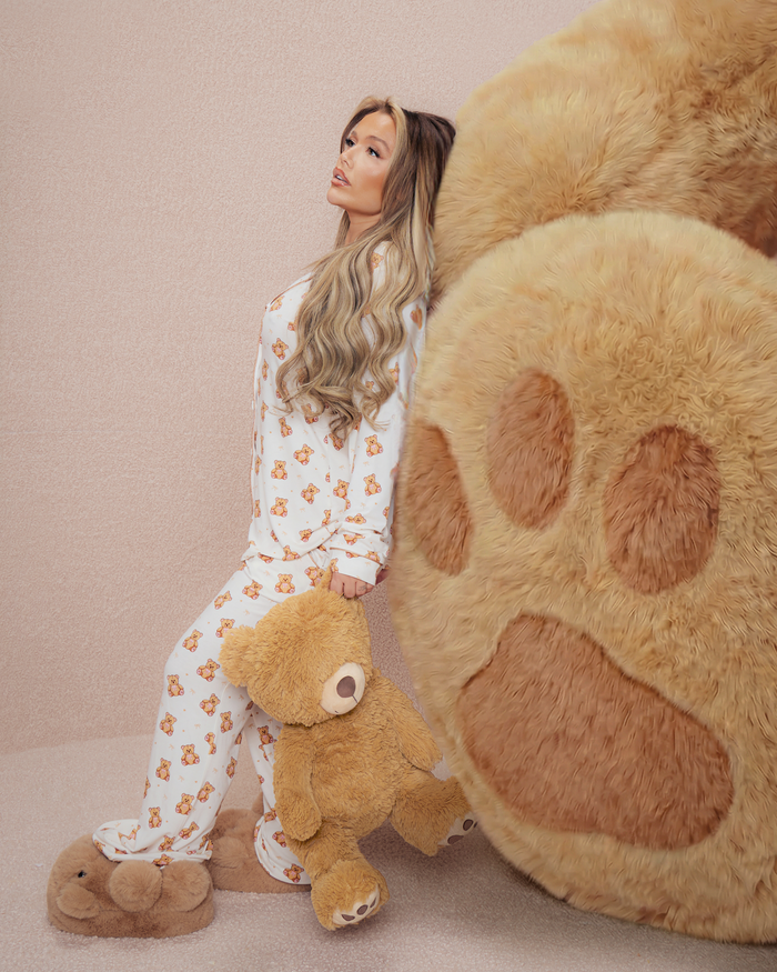 Woman in teddy pyjamas holding a teddy bear next to a large plush bear paw on a beige background